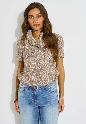 Mujer con cabello castaño largo que lleva una blusa floral de manga corta con cuello con volantes y vaqueros azul claro, de pie sobre un fondo blanco.