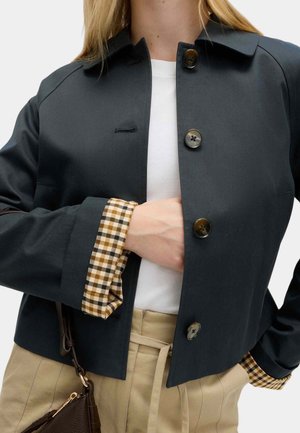 Žena v tmavé zapnuté bundě s kostkovanými manžetami, bílá košile, béžové kalhoty s vysokým pasem, drží hnědou koženou kabelku.