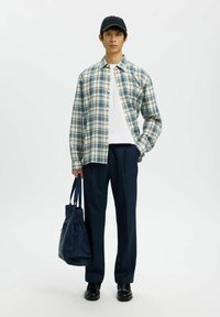 Jeune homme portant une casquette noire, une chemise à carreaux bleue et blanche par-dessus un t-shirt blanc, un pantalon bleu marine, des chaussures noires, tenant un sac fourre-tout bleu marine, debout sur un fond blanc.