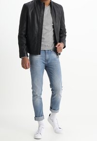 Man in een zwart leren jack over een grijze shirt, lichte blauwe opgerolde jeans, witte sokken en witte sneakers die tegen een witte achtergrond staat.