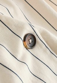Beige stof met marineblauwe verticale strepen; heeft een ronde, donkere knop met vier gaten, die textuur en een subtiele glans toont.