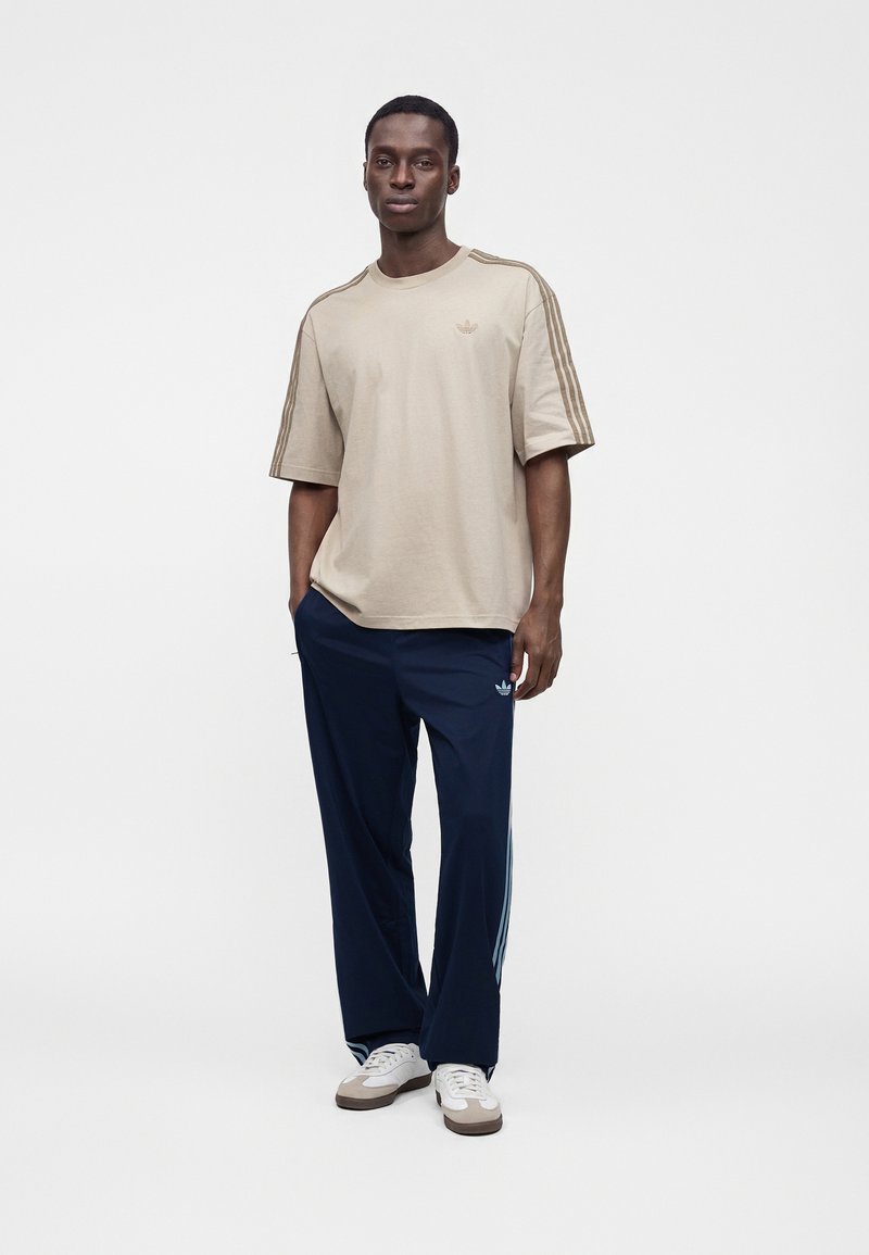 Man wearing beige Adidas t-shirt with stripes on sleeves, navy Adidas track pants, and white sneakers, standing against a plain background.