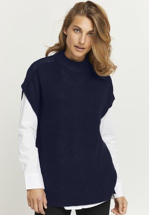 Marineblauer gestrickter Pullover mit hohem Kragen und kurzen Ärmeln, kombiniert mit einem weißen Hemd. Der Pullover hat einen lockeren Schnitt und seitliche Schlitze.