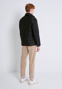 Chaqueta acolchada negra con diseño acolchado, textura suave y cuello alto. Combinada con pantalones de pana color beige y zapatillas blancas.