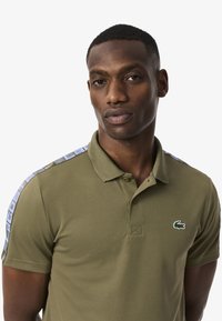 Polo shirt verde oliva con colletto, dotato di un patch con logo e accenti decorati lungo le spalle. Realizzato in tessuto testurizzato.