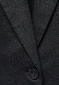 Gros plan d'un blazer en tissu noir texturé avec un revers en encoche et un seul bouton recouvert fermé à l'avant.