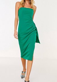 Grön axelbandslös midiklänning i lätt tyg, med draperad sidodel och sidoklipp. Modell som bär mångfärgad mönstrade skor.