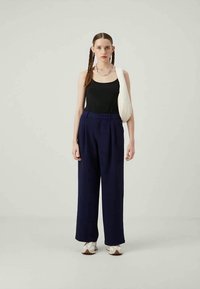 Top preto, calças largas azul-marinho com pregas e bolsa off-white. Modelo com cabelo trançado e calçado em tons neutros. Fundo simples.