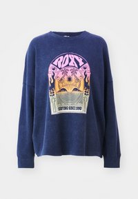 Blaues Waffle-Knit-Sweatshirt mit lässigem Schnitt, ausgestattet mit einem bunten Grafikdesign mit der Aufschrift "ROXY Surfing Since 1990" auf der Vorderseite.
