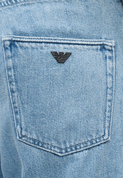 Φωτεινή μπλε τζιν πίσω τσέπη με ραμμένο περίγραμμα και ένα μικρό μαύρο μεταλλικό τριγωνικό λογότυπο στο κέντρο κοντά στην κορυφή.