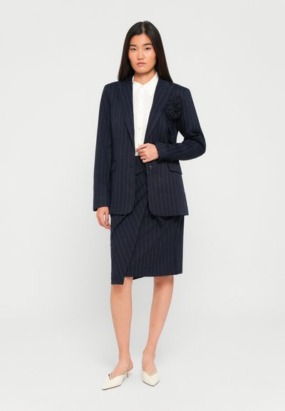 Kvinne i en marineblå stripete blazer med blomsterdetalj, matchende omslagskjørt, hvit skjorte med knapper og hvite lave hælsko mot en hvit bakgrunn.