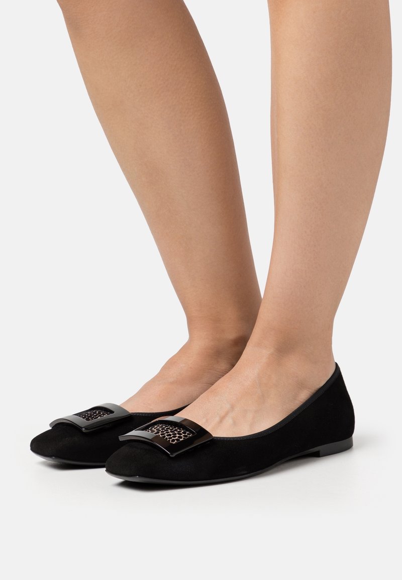 Pieds portant des ballerines en daim noir avec une boucle carrée noire et un détail imprimé léopard sur fond blanc.