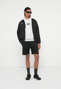 Chaqueta bomber negra con costuras blancas, sudadera gris con logo, pantalones cortos negros con logo rojo, calcetas oscuras y calzado negro.