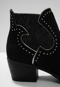 Bottine en suède noir avec un panneau textile à côtes, des accents cloutés, des bords festonnés et un chunky talon en bois.
