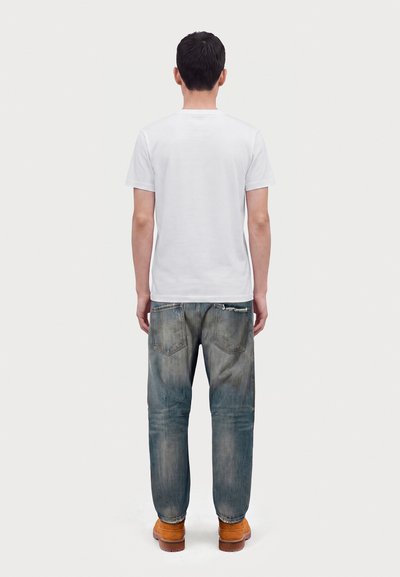 Homme debout de dos, portant un t-shirt blanc uni, un jean bleu délavé et des bottes marron sur un fond clair uni.