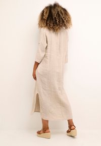 Linen dress in beige with three-quarter sleeves, a back slit, and a loose fit. Paired with brown wedge sandals.