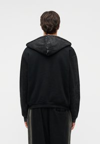 Άνδρας που φοράει μαύρο φουτερ με κουκούλα και σκούρο παντελόνι, στέκεται με την πλάτη στην κάμερα μπροστά σε απλό φωτεινό φόντο.