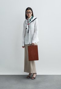 Bijela lanena košulja s vezanom maramom, bež dugačka suknja i crne sandale s potplatom. Drži smešnu kožnu torbu s ugraviranim detaljima.