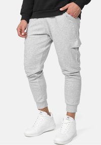 Mies, joka käyttää vaaleanharmaita jogger-housuja, joissa on sivutasku, sekä valkoisia korkeita sneaker-kenkiä, käsi taskussa, taustana yksinkertainen tausta.