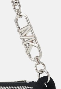Srebrny metalowy zamek z logo "A|X" łączy czarny materiałowy pasek i zamek błyskawiczny na torbie z ćwiekami na białym tle.