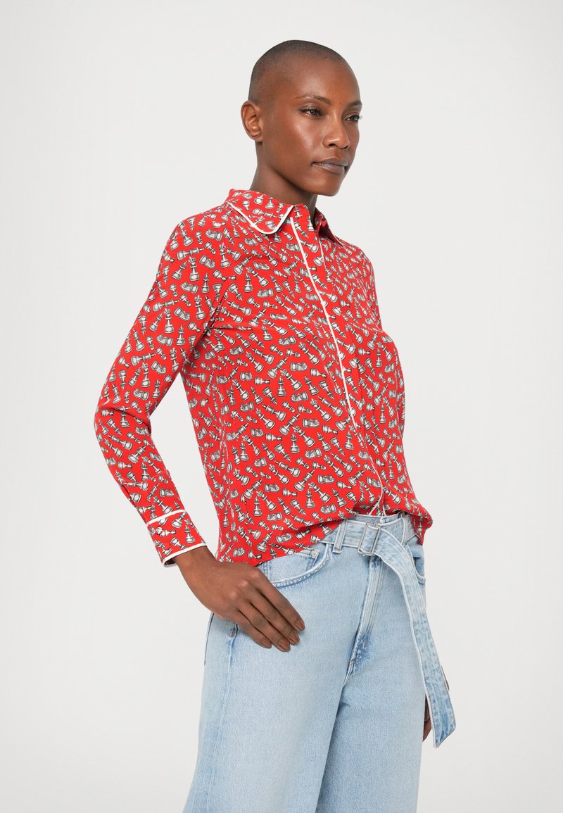 Camisa roja de botones con detalles en blanco y un patrón juguetón de pequeños diseños en gris y blanco. Mangas largas combinadas con jeans anchos de tono azul claro.