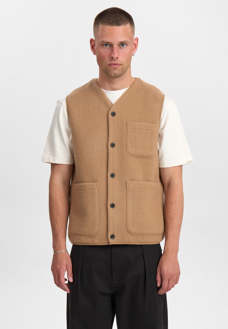 Gilet di lana kaki con scollo a V, tasche frontali e bottoni neri. Indossato sopra una camicia bianca, abbinato a pantaloni neri. Tessuto liscio.