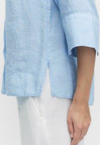 Ljusblå linneskjorta med avslappnad passform, ärmlängd till armbågen och sidslitsar i fållen. Slät textur med minimala sömmar.