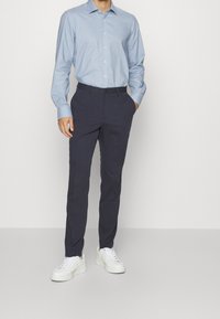 Uomo che indossa una camicia azzurro chiaro con bottoni, pantaloni slim fit blu scuro e sneakers bianche, in piedi con una mano in tasca.