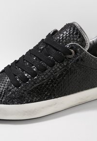 Sneaker nera con motivo reptilico testurizzato, lacci neri, dettagli argentati e una suola in gomma bianca. Leggeri segni di usura visibili sul bordo della suola.