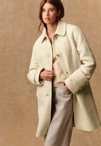 Vrouw in een crèmekleurige getextureerde jas over een beige gebreide trui en lichtgrijze broek, poseert met één hand in de zak tegen een neutrale achtergrond.