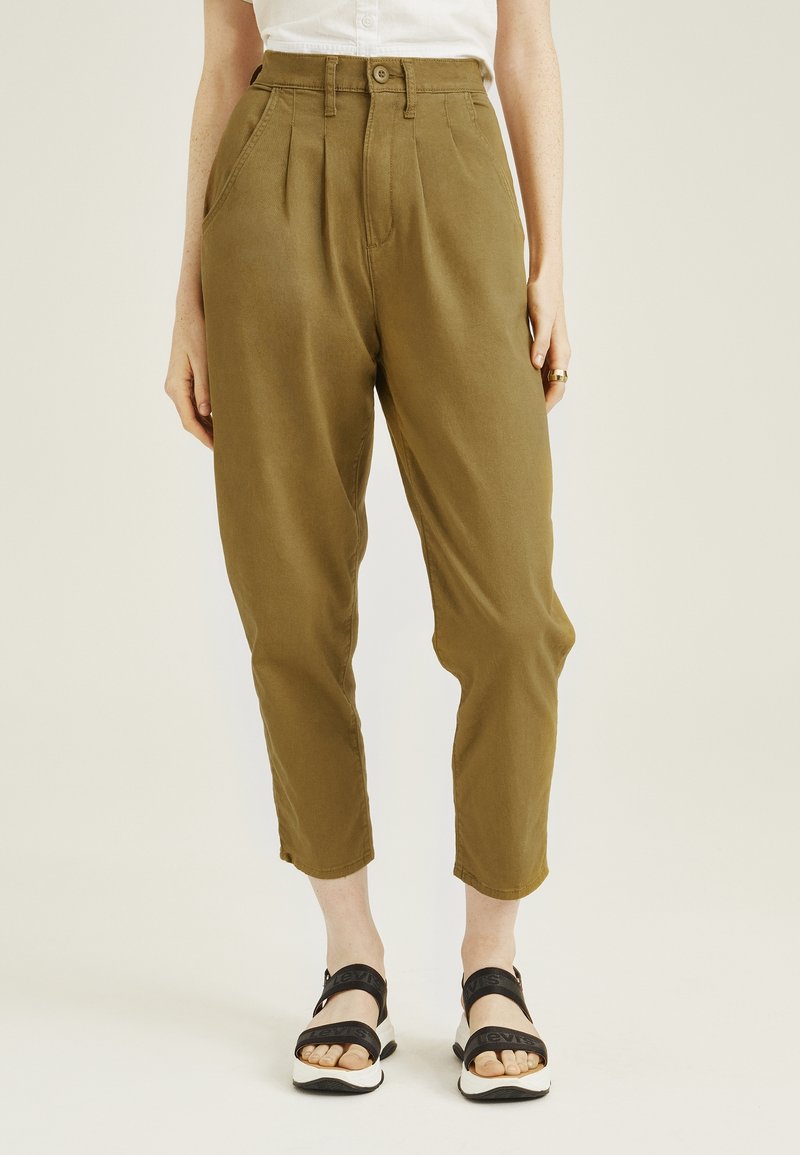 Pantalon taille haute vert olive avec plis, jambes fuselées et poches latérales. Associé à des sandales noires à lanières sur un fond neutre.
