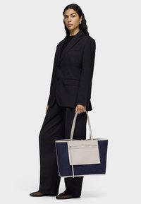Large tote bag featuring a navy fabric body with light gray leather accents, rectangular shape, dual handles, and front zip pocket.