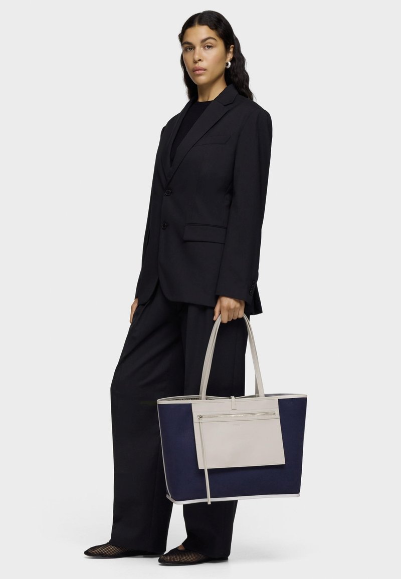 Large tote bag featuring a navy fabric body with light gray leather accents, rectangular shape, dual handles, and front zip pocket.
