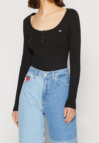 Schwarzer Langarm-Body mit Rundhalsausschnitt und einer dreiknöpfigen Knopfleiste. Kombiniert mit hellblauen Patchwork-Hosen mit hoher Taille und Taschen.