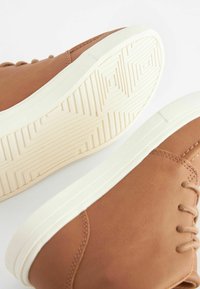Brune lædersneakers med hvide gummisåler, prydet med teksturerede mønstre, en rund tå og tynde snørehuller. Glat finish.