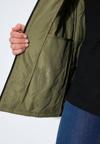 Interno della giacca verde oliva con tessuto leggero e liscio. Presenta una tasca e il testo "idrorepellente" stampato all'interno. Design con zip.