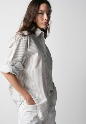 Jonge vrouw met lang donker haar, dragen een losse, opgerolde gestreepte blouse en witte broek, staand met handen in de zakken tegen een effen achtergrond.