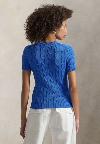 Πλεκτό πουλόβερ με κοντά μανίκια σε φωτεινό μπλε χρώμα, με σχέδιο κοτσίδας, εφαρμοστή γραμμή και στρογγυλή λαιμόκοψη, φορεμένο με ανοιχτόχρωμο παντελόνι.