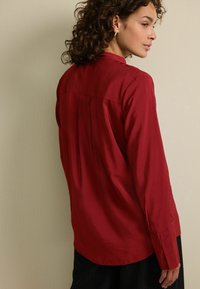 Červená košile s dlouhým rukávem s hladkým povrchem, detailem záhybu na zádech a knoflíkovými manžetami. Límec je tradiční s jemným leskem.