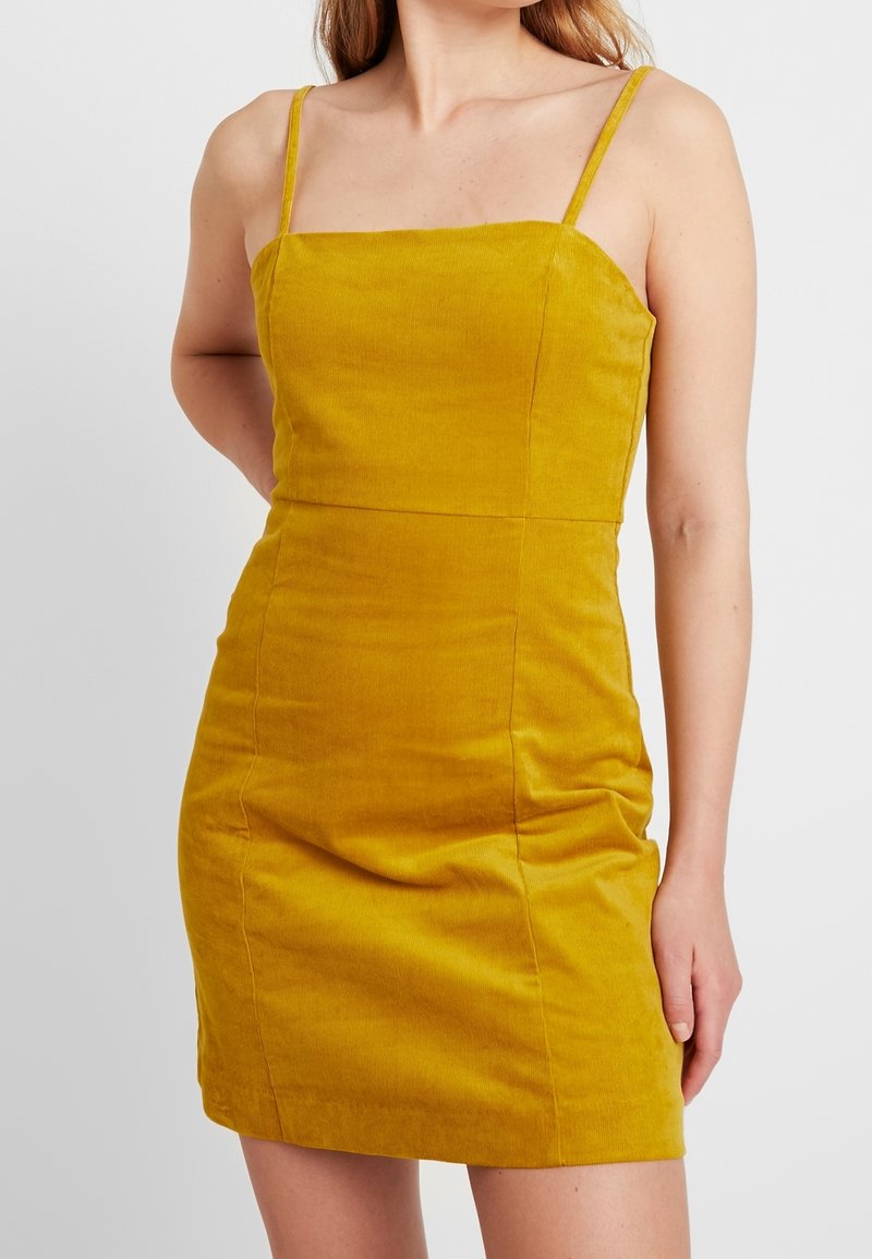 Femme portant une mini-robe sans manches en velours côtelé jaune moutarde avec de fines bretelles, debout devant un fond blanc uni.