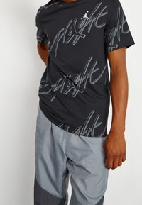 Homme portant un t-shirt noir Jordan Flight avec un texte gris et un pantalon gris clair, se tenant devant un fond uni.