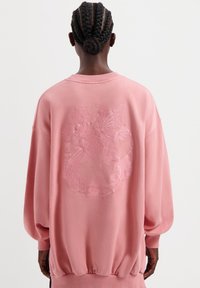 Roze sweatshirt met een ronde hals, lange blousonmouwen en een geribbelde onderrand. Heeft een reliëf bloemenontwerp op de achterkant.
