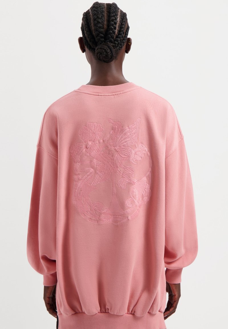 Roze sweatshirt met een ronde hals, lange blousonmouwen en een geribbelde onderrand. Heeft een reliëf bloemenontwerp op de achterkant.