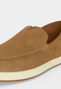 Chaussure slip-on en daim marron avec semelle blanche et coutures apparentes sur la tige, vue en angle montrant le dessus et le côté.