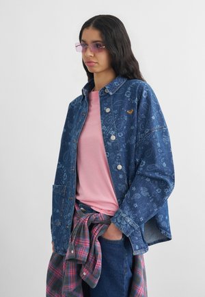 Mladá žena v kvetinovej rifľovej bunde cez ružové tričko s károvanou košeľou previazanou v páse a fialovozafarbenými slnečnými okuliarmi stojaca pred bielym pozadím.