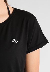 Černé tričko s krátkým rukávem vyrobené z hladké tkaniny, s jemným bílým logem na levé straně. Jednoduchý a ležérní design.