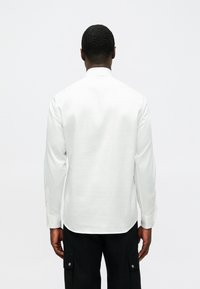 Camisa branca de mangas longas feita de tecido leve, com colarinho clássico e punhos com botões, apresentada pela parte de trás sem padrões visíveis.