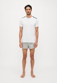 T-shirt bianca a maniche corte con dettagli neri sulle spalle, abbinata a pantaloni corti grigi chiari. A piedi nudi, in piedi su uno sfondo neutro.