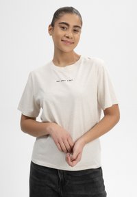 Cremefarbendes Baumwoll-T-Shirt mit kurzen Ärmeln, das vorne mit dem schwarzen Drucktext "EINEN APFEL AM TAG" versehen ist. Kombiniert mit dunklen Jeans.