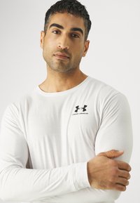 Bijela dugorosna majica napravljena od laganog materijala, s crnim Under Armour logotipom na lijevoj strani grudi. Glatka tekstura s uskim dizajnom.